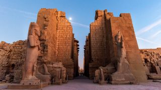 tall egyptian ruins with tall figures and walls with a blue sky and sun in the background.