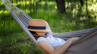 Woman resting in a hammock in a summer garden covering her face with a straw hat.