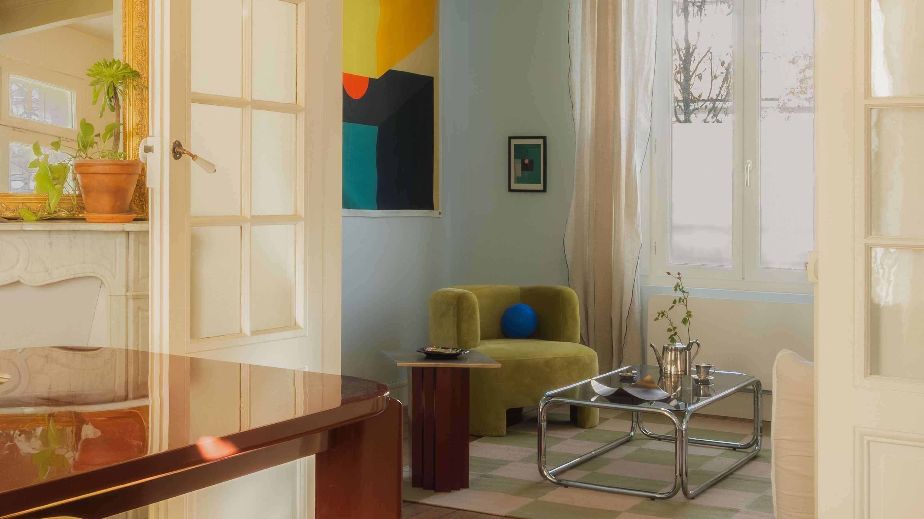 A living room with a lime-toned armchair, abstract colorful artwork, pale blue walls, a tubular chrome and glass coffee table, and white-washed wooden detailing.