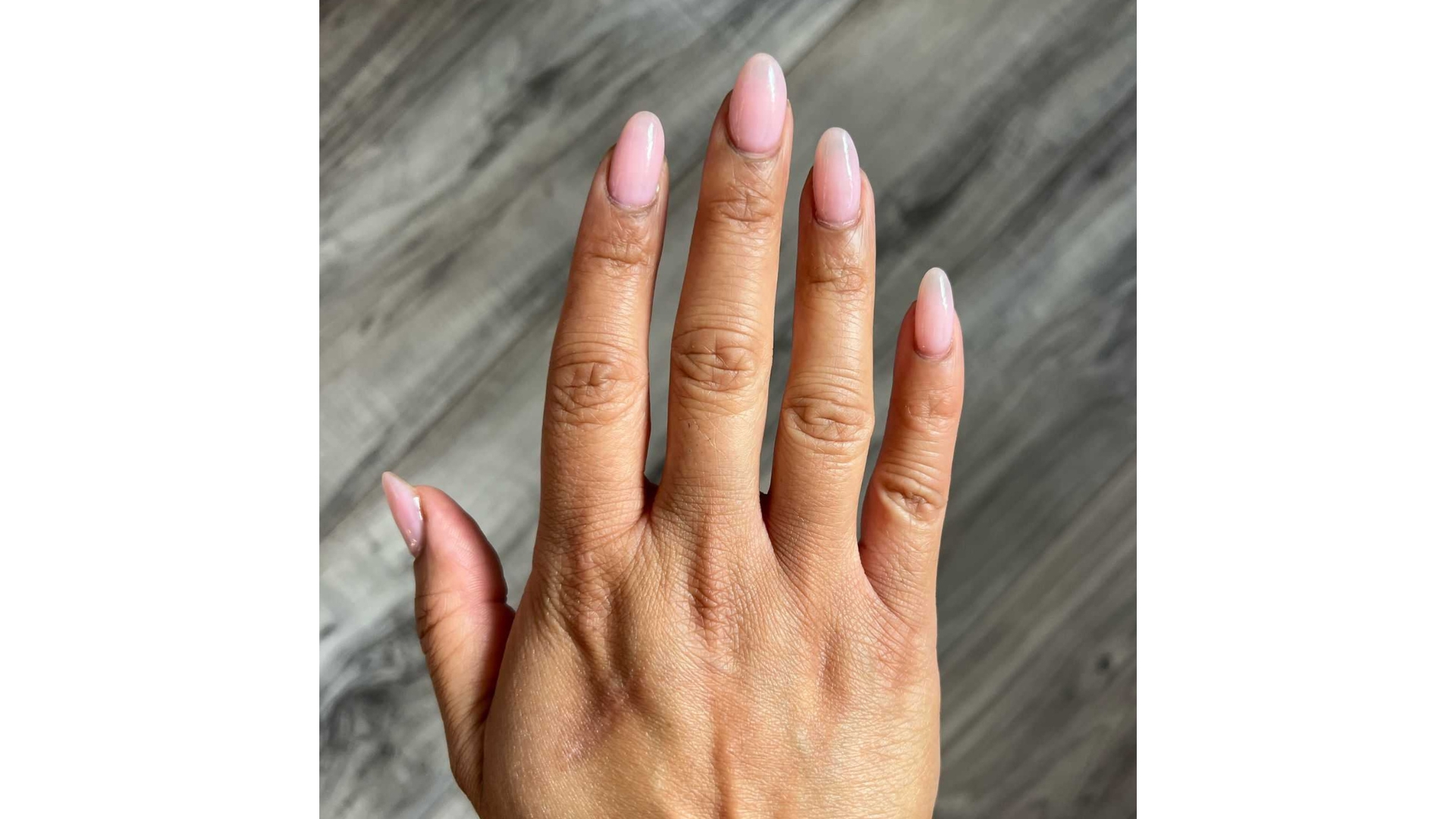 A close-up of Digital Ecommerce Editor, Aleesha Badkar's hand, which features a candy-pink almond manicure, pictured in front of a grey wood flooring background