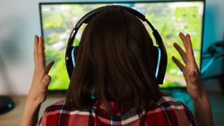 A woman pictured from behind who is holding her hands out in frustration and wearing a headset. She is seated at a table with a desktop monitor in the background.