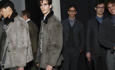 Six male models wearing looks from Giorgio Armani's collection. Two are wearing dark suits, blue shirts, waistcoats and glasses. A third model is wearing a brown suit, a fourth model is wearing a dark shirt and grey trousers and the final two models are wearing grey and black short fur jackets