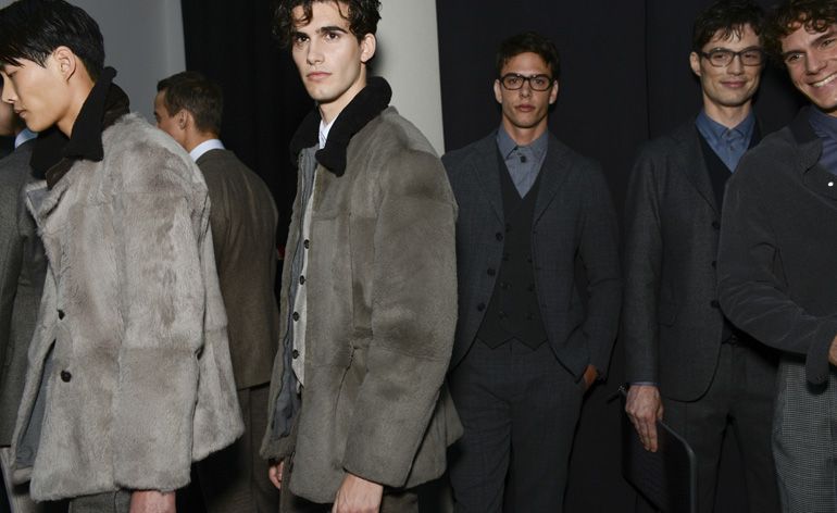 Six male models wearing looks from Giorgio Armani&#039;s collection. Two are wearing dark suits, blue shirts, waistcoats and glasses. A third model is wearing a brown suit, a fourth model is wearing a dark shirt and grey trousers and the final two models are wearing grey and black short fur jackets