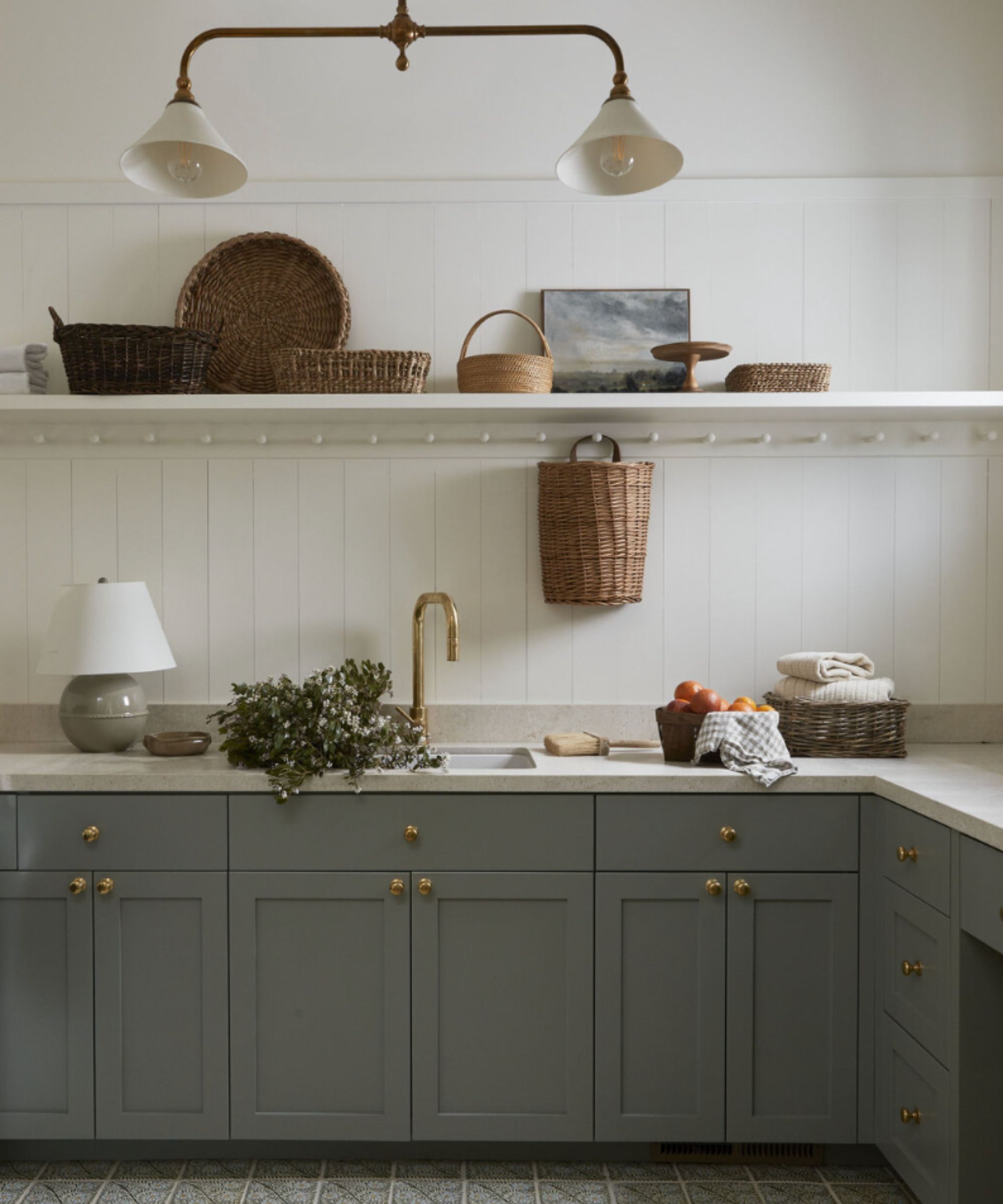 Kitchen with white panelled walls, light gray cabinetry, shelf above counter decorated with baskets and artwork, counter with sink decorated with baskets and lamp