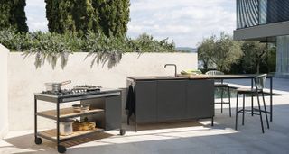 Outdoor kitchen setup pool side with a kitchen sink with breakfast table module and 2 kitchen stools, cooking module with wheels with a view of the mountains and green plants/trees in the background