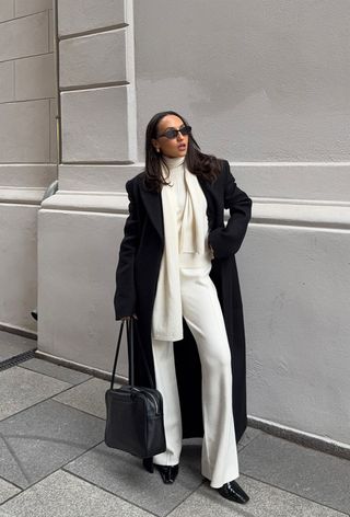 A woman standing outside wearing a long black coat over a white sweater with matching knit pants styled with black kitten-heel boots and a black bag.