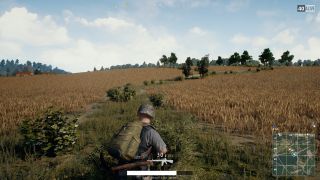 Over the shoulder PUBG gameplay with tactically armed protagonist looking at typically beige PUBG field of wheat with trees in background