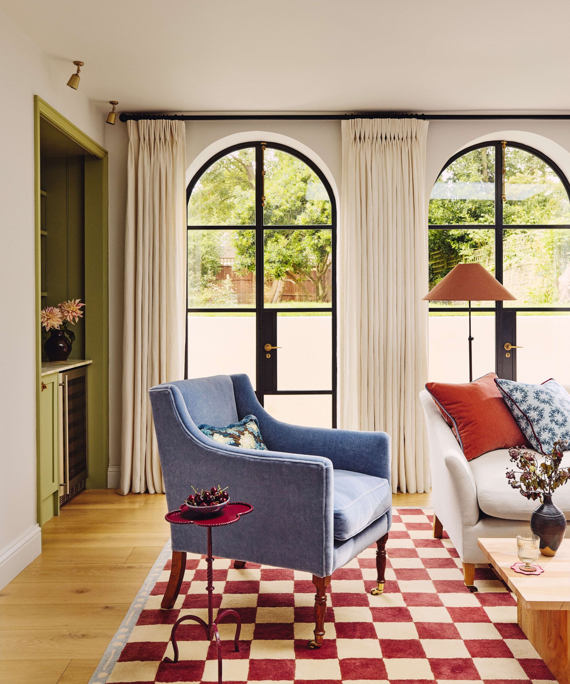A living room featuring large arched black-framed windows and long cream curtains. A plush blue armchair and a white sofa with colorful cushions sit on a bold red-and-cream checkered rug. There is built in cabinetry painted in an olive green on the left hand wall.