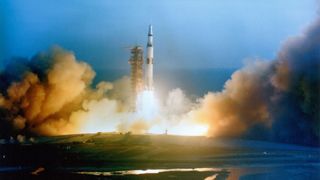 a saturn v rocket lifts off into the blue sky as seen from a distance