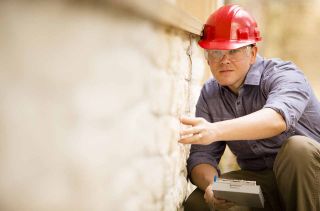 Repairman, building inspector, exterminator, engineer, insurance adjuster, or other blue collar worker examines a building/home exterior wall.He wears a red hard hat and clear safety glasses