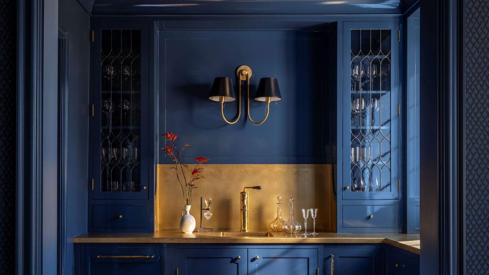 Dark blue painted home bar with built-in glass cabinetry, gold backsplash and countertops, and a large sconce in the centre 