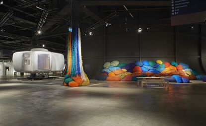 A museum-like room with colorful tufts of fabric of fabric in the background mounted on a black-painted curve wall and some hanging off the ceiling. Oak coloured long table and chair. 