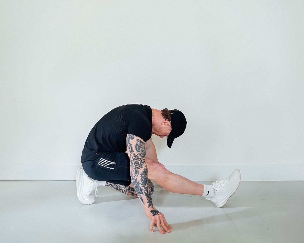Coach Corey Perkins performs a calf stretch. His left leg is bent, with his toes and knee on the floor and his buttock resting on his heel. His right leg is held out straight in front of him. His back is curled over and his head points down. His hands rest either side of him on the floor.