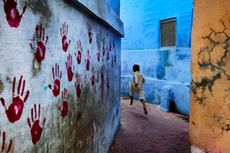 Boy in mid-flight’, Jodhpur, India, by Steve McCurry, 2007. A boy running down an alley with red hand prints on the wall.