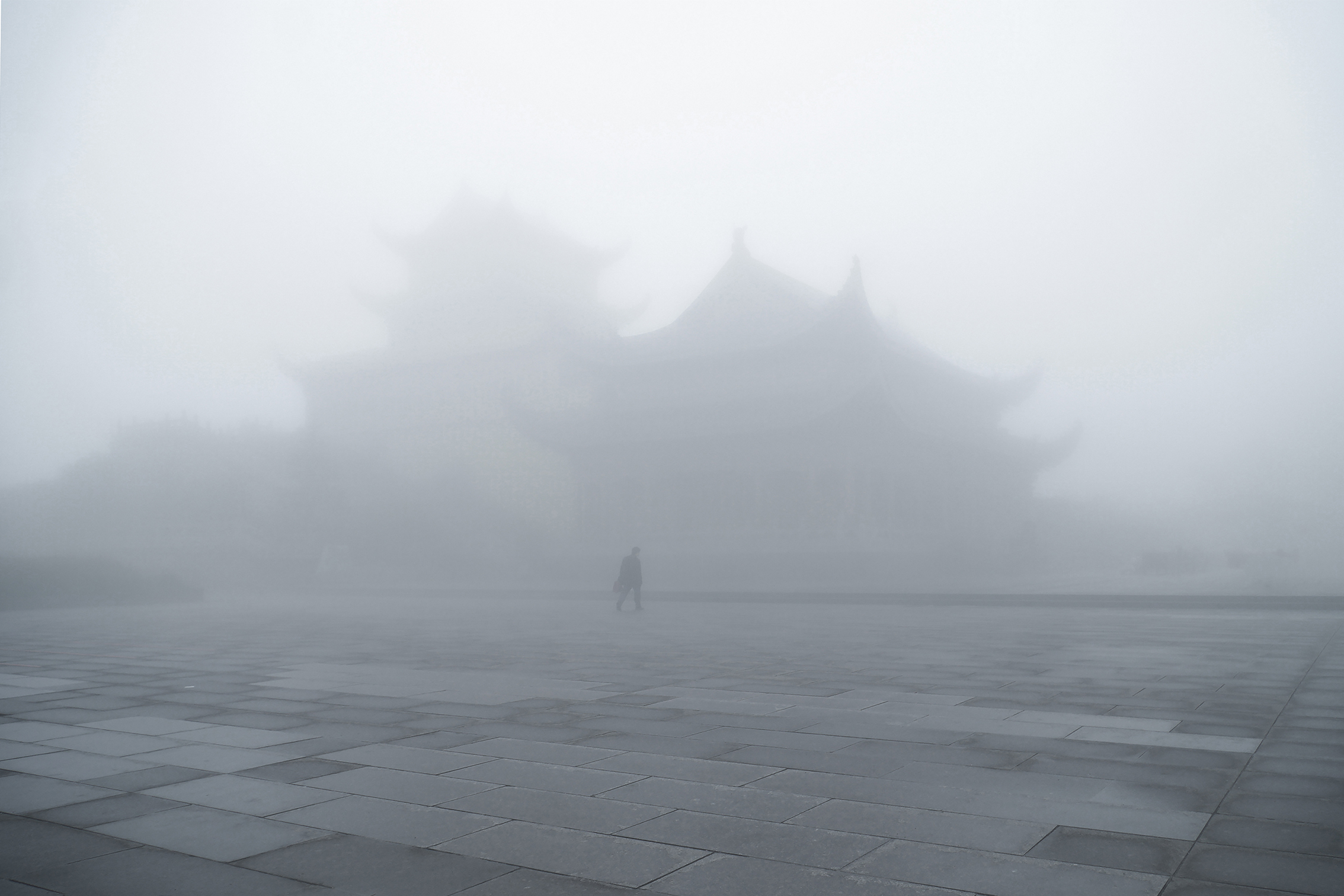 A lone figure walks through thick fog obscuring traditional Asian-style buildings, creating a mysterious and tranquil atmosphere on an empty stone plaza