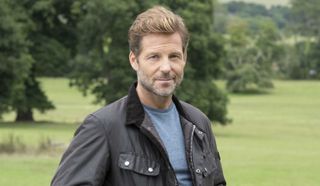 Jamie Bamber as Archie Hughes in Beyond Paradise, wearing a dark jacket and standing in a field