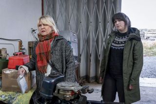 Calder (Ashley Jensen) and Tosh (Alison O'Donnell) stand inside a warehouse or workshop of some kind - there is a large industrial concertina-style door behind them that's half open, and various items like a small bedside cabinet and an outboard motor scattered around in need of repair