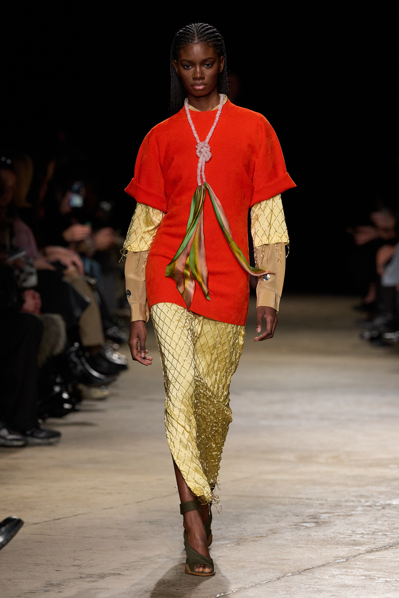 A model on Zankov's Fall 2026 runway wearing an electric orange persimmon colored short sleeve sweater, butter yellow midi dress, chainmail top layer, and brown long-sleeve shirt.