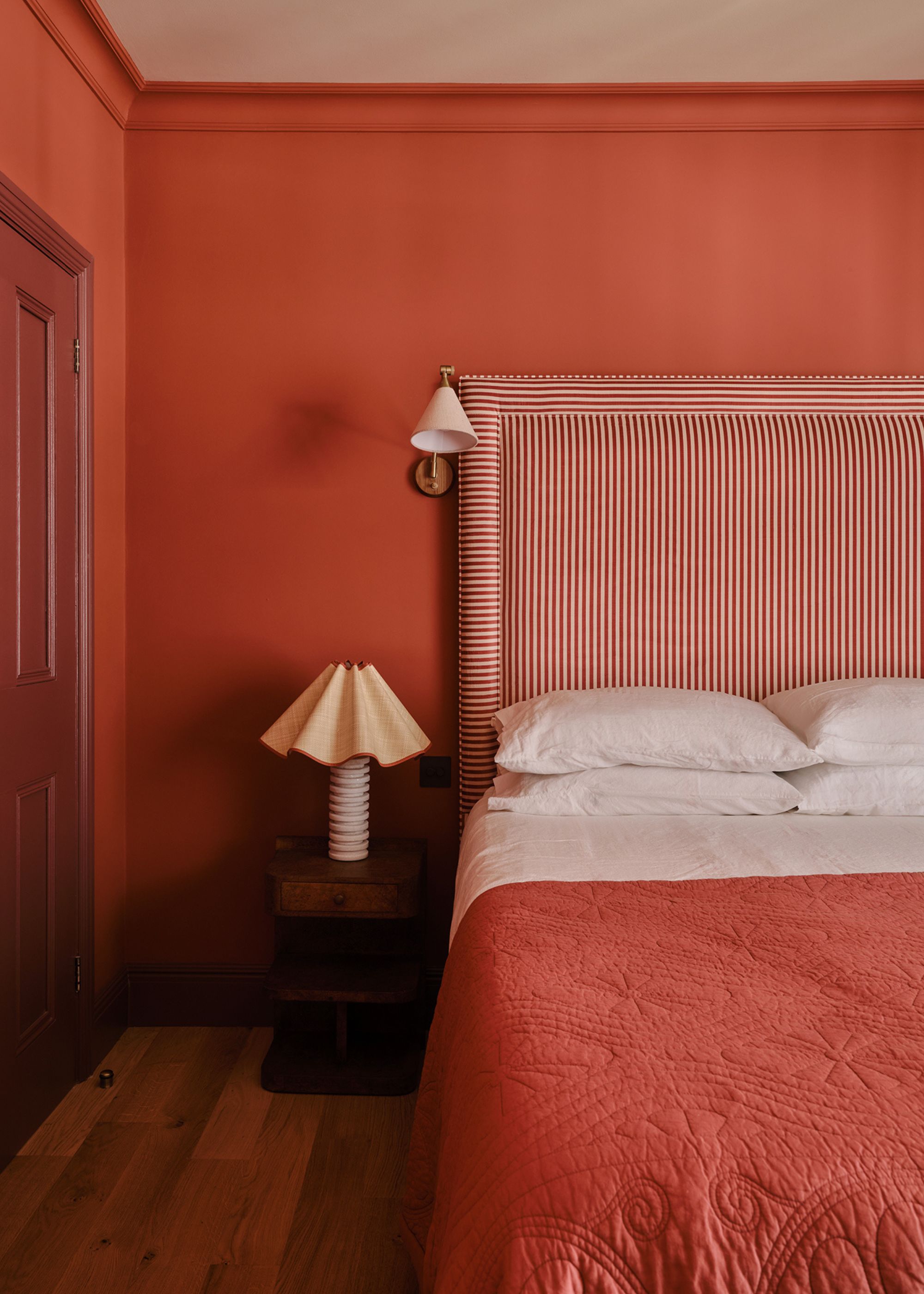A bedroom painted a terracotta red with a red and white striped headboard, white and brick red bedding, and a nightstand with a wavy lampshade.