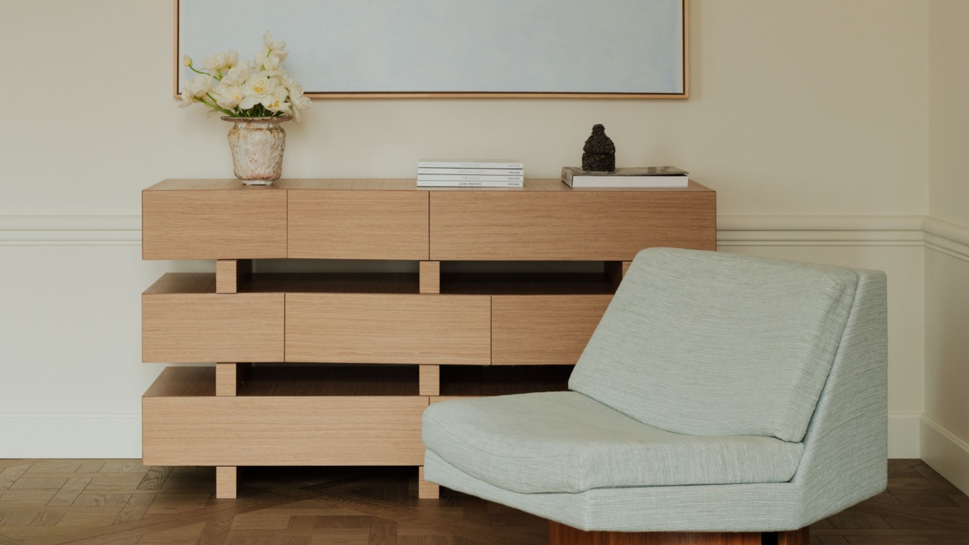 A living room corner with a wood sideboard, a vase of flowers, a stack of books, and a low-slung pale blue chair
