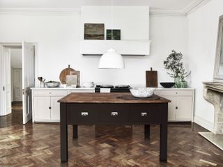 a stunning white kitchen with darker accents, including a dark table in the middle with a wooden top, and a marble fireplace to the right, with wood parquet flooring