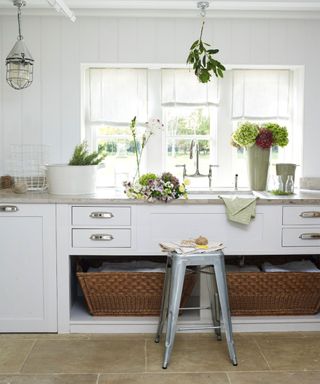 White cabinetry with drawers and storage baskets and stone countertops in front of a window illustrating how to organize a laundry room.