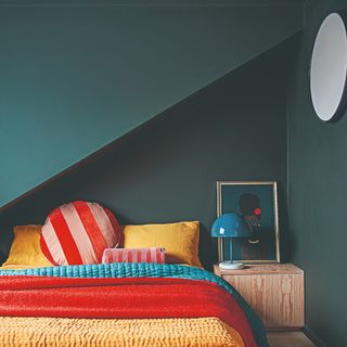 A teal-painted colour-drenched bedroom with colourful bedding on the bed and a matching table lamp on the bedside table