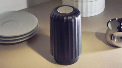 Image of a black, ribbed coffee capsule crusher on a beige countertop. There is a stack of small white plates behind it and a silver dish in view. The image is grainy like film, and there are shadows from the items on the countertop.