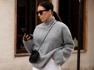 British fashion influencer Jessica Skye poses with black sunglasses, a gray mock neck turtleneck sweater, silver chunky chain necklace, black crossbody mini bag, and white jeans.