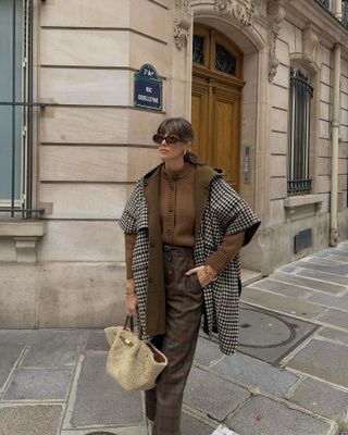 French influencer wears brown cardigan, plaid winter coat, plaid trousers and sherpa handbag, standing on Paris street.