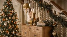 A foyer with a white banister with green garland, a wood sideboard with an inclusion candle, a brassy lamp, and tapers by a Christmas tree in autumnal baubles