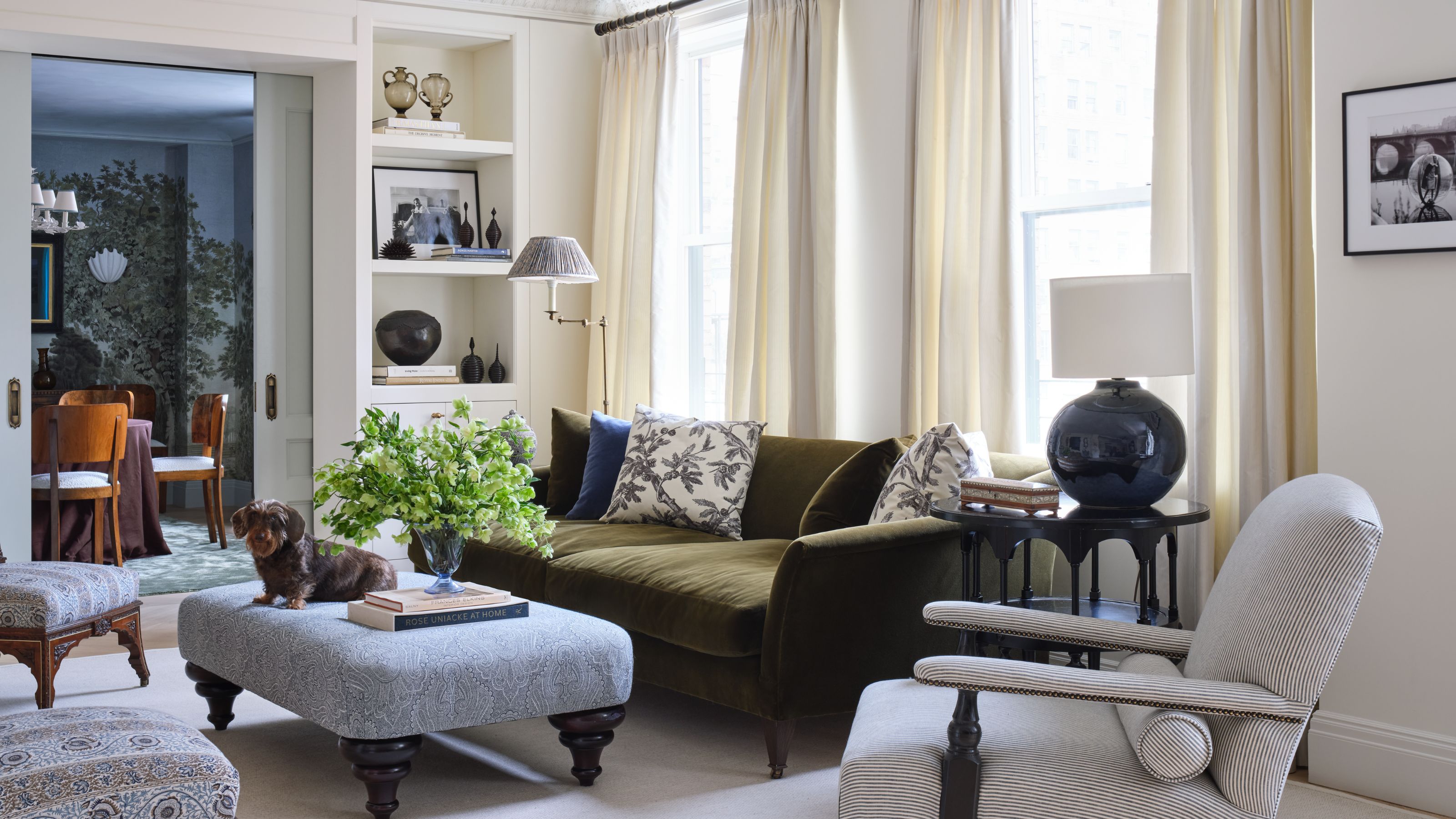 a large formal living room in New York with elegant moldings, neutral walls, doorway bookcases, olive green velvet couch, small accent chairs and an ottoman