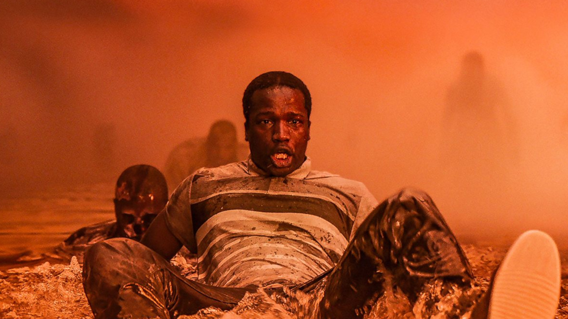 Sope Dirisu as Bol sat in water in a room covered in orange light during the horror movie, His House.