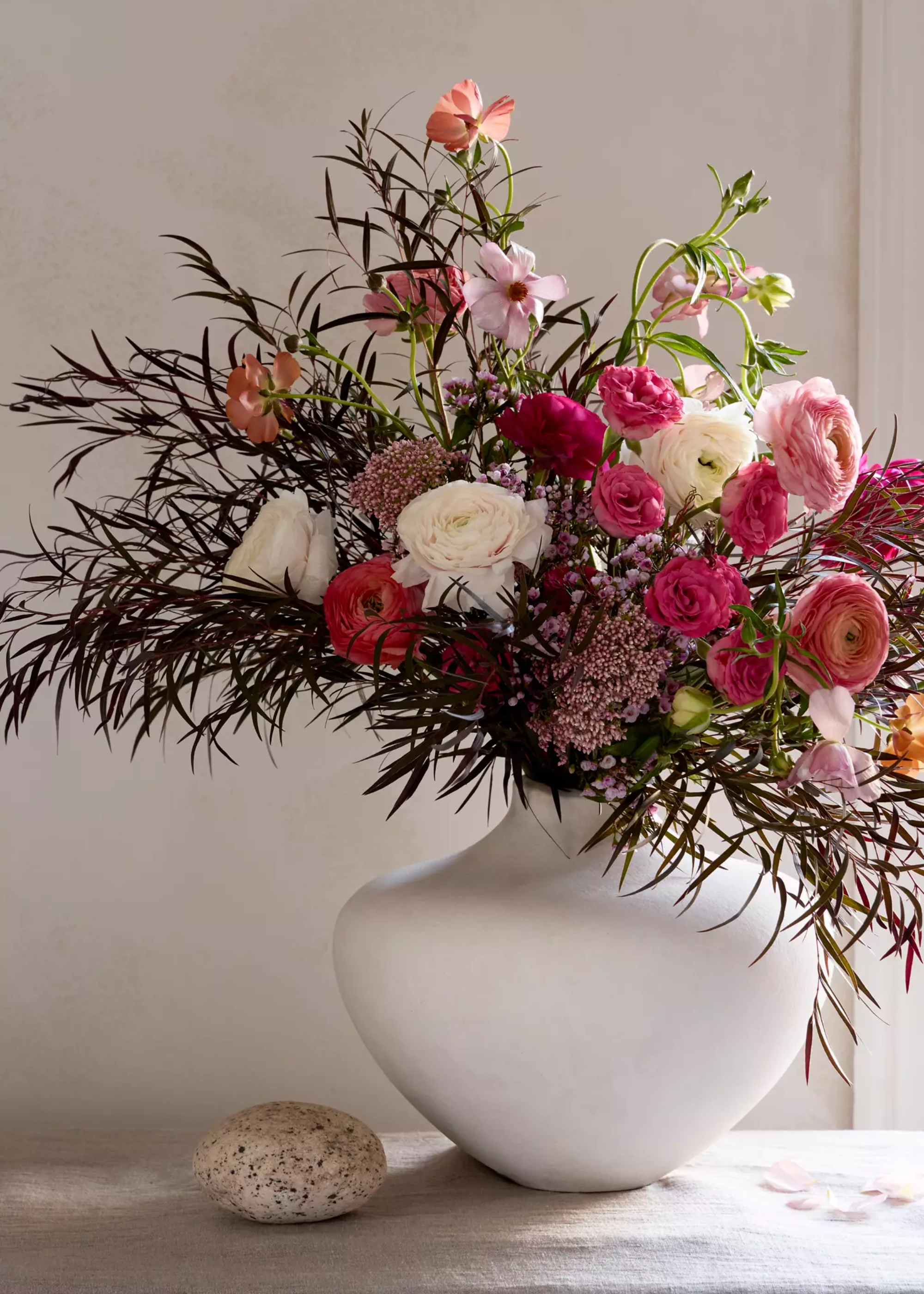 A vase of roses and ranunculus with feather foliage in a white vase from Terrain