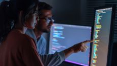 Two cybersecurity workers looking at a portrait monitor showing code. One points at the screen, which lights up their faces in the dark room, while in the background a landscape monitor shows more code.