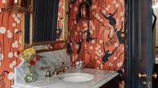 A small marble sink and backsplash in a small bathroom with terracotta wallpaper showing stripes, round white anterns and monkeys. The hardware and mirror is brass and the wooden door is painted dark blue