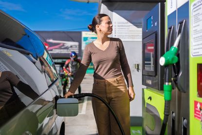 Woman refuels her car at a gas station on a sunny day, pondering rising gas prices.