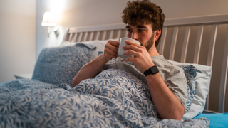 A man lying in bed propped up by pillows drinking a cup of tea from a mug