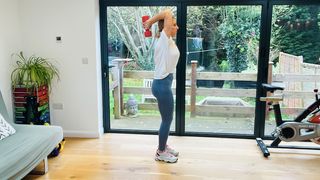 Maddy Biddulph performs a triceps hold at home in her living room. She wears leggings, a t-shirt and sneakers. She is standing and holding a dumbbell in her hands behind her head. Her elbows are bent and held close to her head. Behind her we see glass sliding doors leading to a garden and an exercise bike.