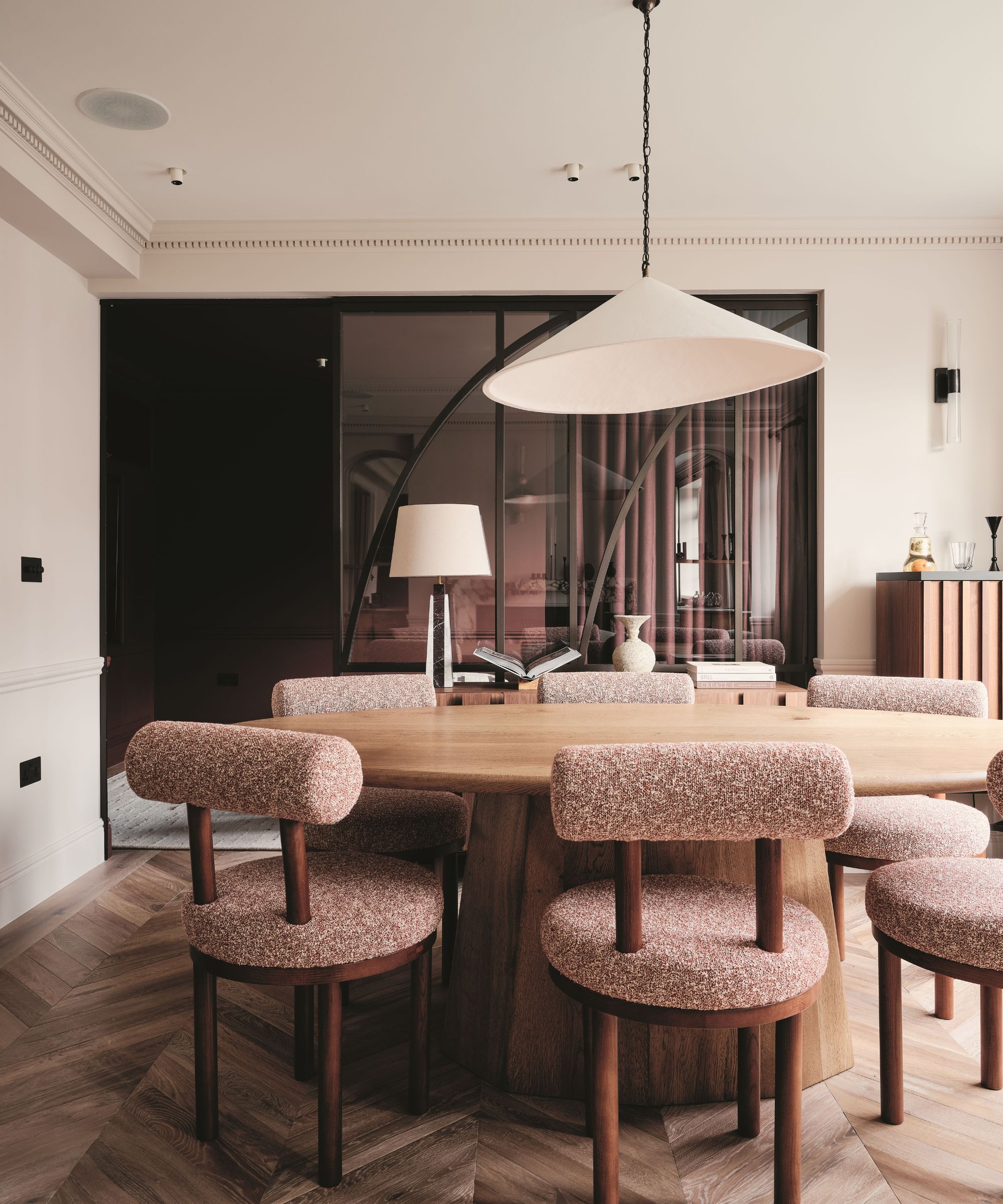 Oval oak dining table with mauve boucl&amp;eacute; chairs under a large conical pendant. A curved glass partition and chevron wood floors complete the modern, warm interior.
