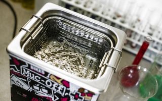 A shiny silver chain sits in a water bath within a cubic cleaner covered in stickers