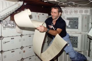 a man in a blue shirt holds a large scroll of teleprinter copy while floating aboard a spacecraft