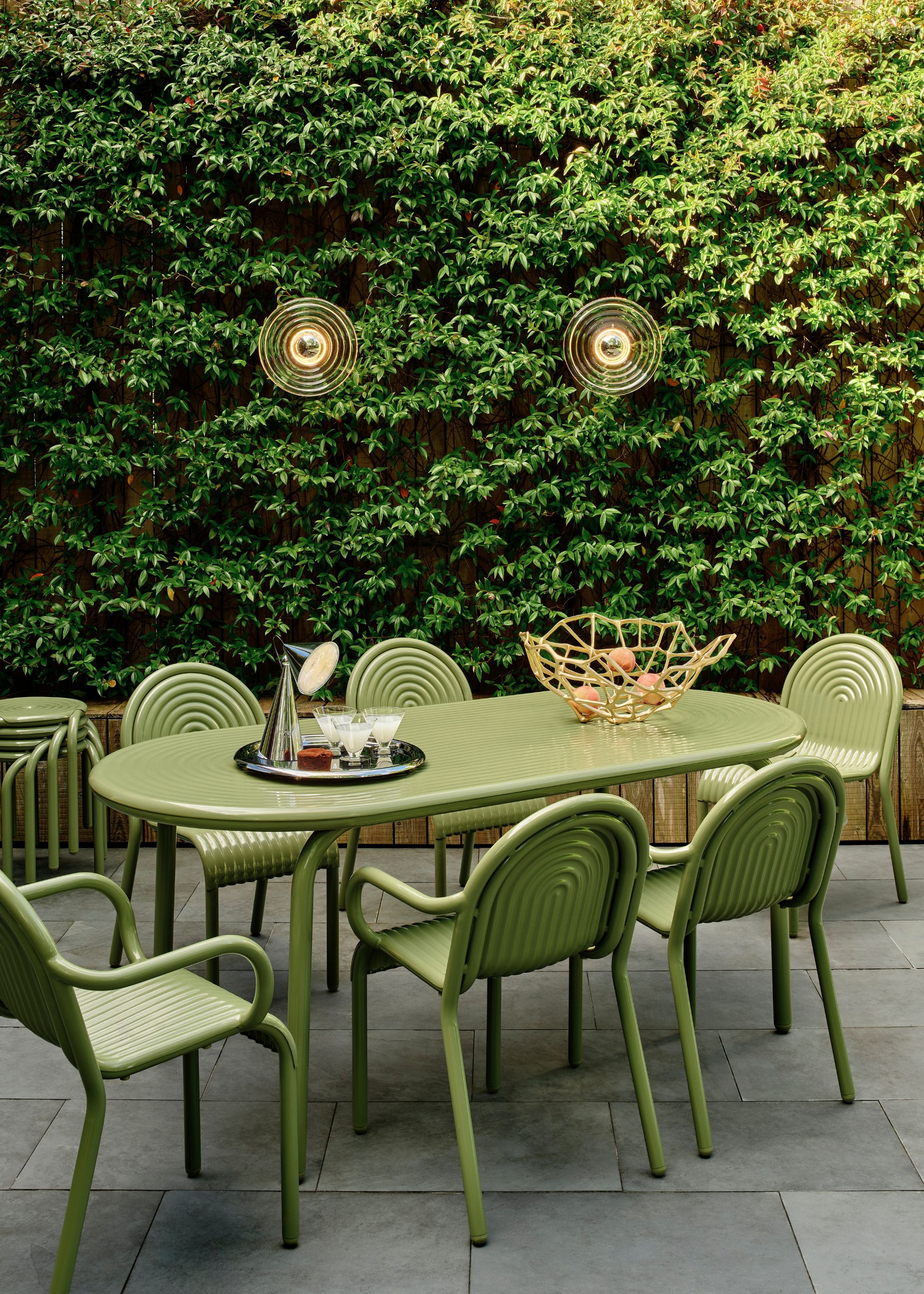 A green colored table and chairs with a silver tray and silver lamp on top. There is also a netted gold basket on top of the table with fruits inside