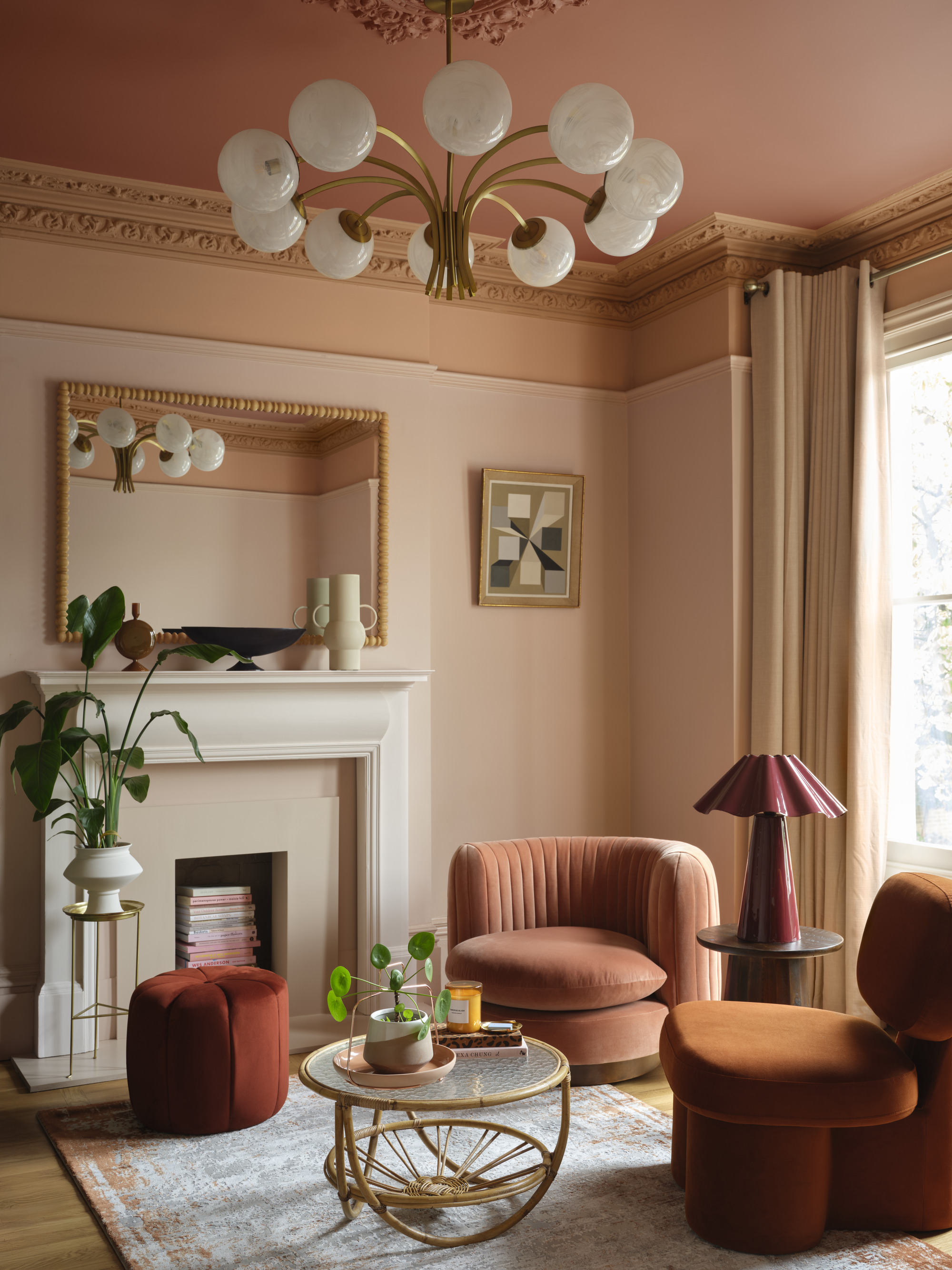 A living room painting in a color capping technique using varying shades of peach and pink paint colors. There is crown molding around the ceiling and a white bulb light fixture. There are two pink and orange, velvet accent chairs and a fire place in the room.