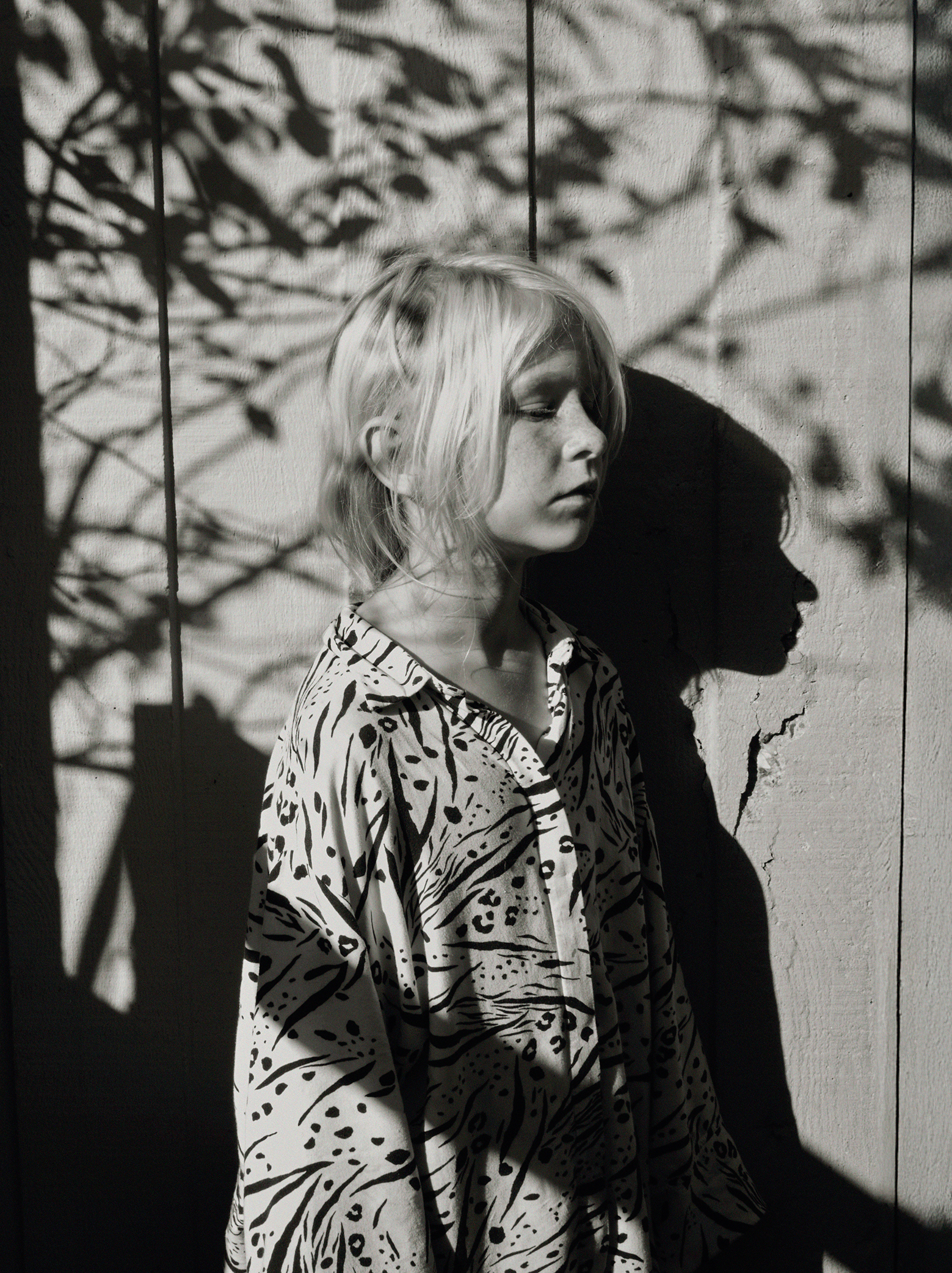 Young person with light hair in patterned shirt stands against a textured wall. Shadows from leaves fall across both. The mood is serene and contemplative