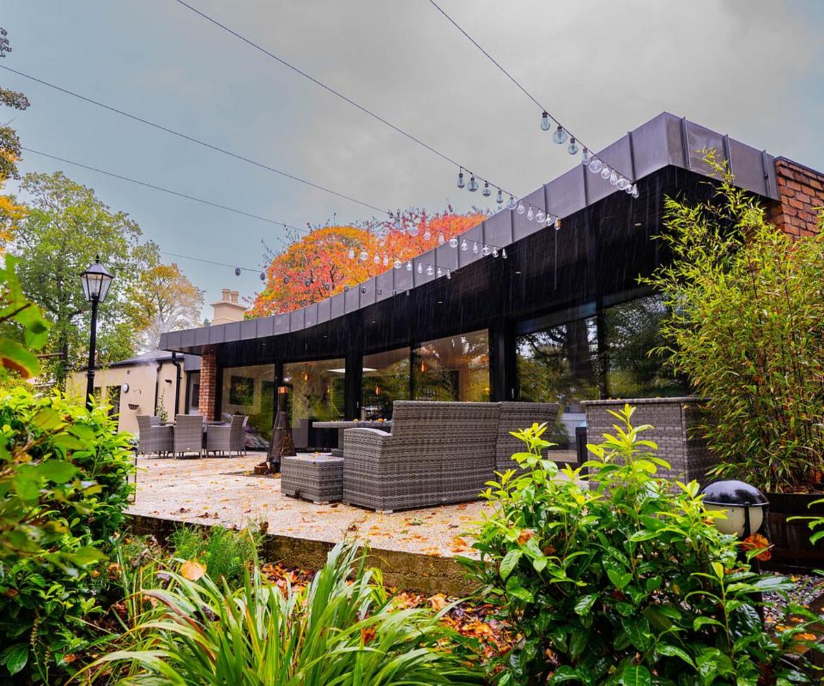 one side of extension showing glass facade with roof overhang in zinc cladding, leading to patio area with seating and onto garden