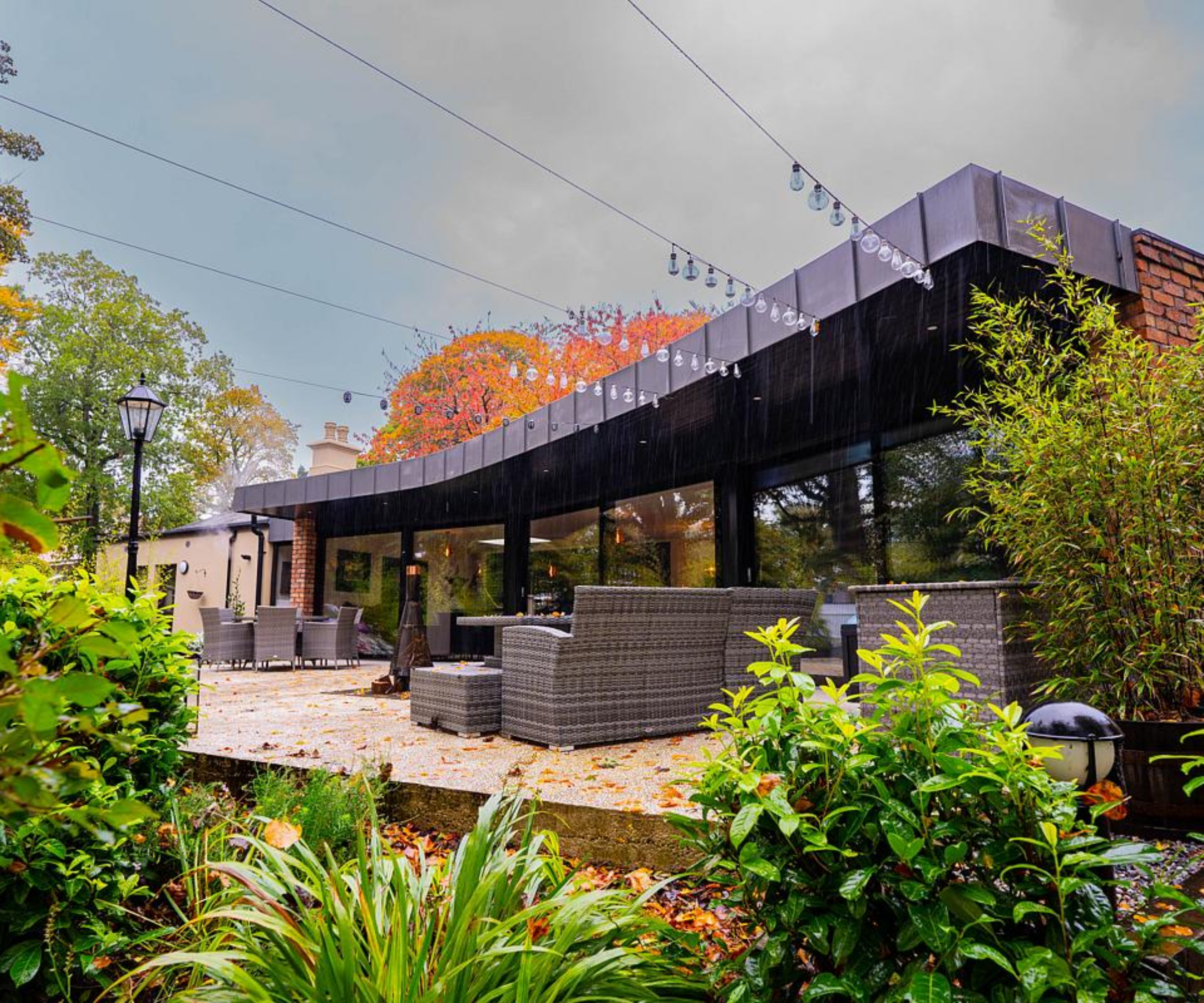 one side of extension showing glass facade with roof overhang in zinc cladding, leading to patio area with seating and onto garden