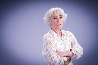 A posed shot of Charlie Hungerford (Zoë Wanamaker). She is wearing a white dress with an irregular pattern of short horizontal blue lines repeated across it, and a belt around her waist. She has her arms folded and is looking off to the side, as though concerned.