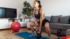 woman in a wide squat position holding a kettlebell with both hands in between her legs. she's on a blue exercise mat in a living room with a red exercise ball visible