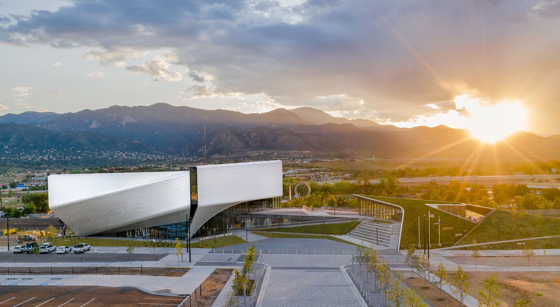 US Olympic & Paralympic Museum is a modern construction, with an asymmetrical metal roof. It's surrounded pa a parking lot, and green surfaces. In the distance, we see mountains and the sun setting behind them.